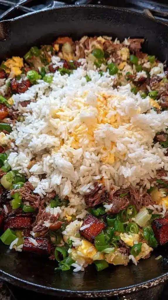 Vertical close-up of a cast-iron skillet with brisket fried rice; a mound of day-old white rice is added to sizzling brisket, scallions, and scrambled egg.
