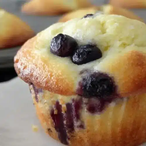 Close-up of a golden blueberry muffin made with cake mix, bursting with juicy blueberries; muffin tin with more muffins blurred in the background.
