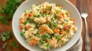 Bowl of creamy crockpot chicken and rice for kids with peas, carrots, and parsley on a white plate.