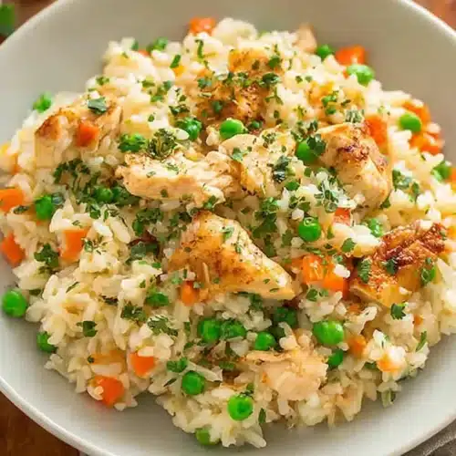 Bowl of creamy crockpot chicken and rice for kids with peas, carrots, and parsley on a white plate.