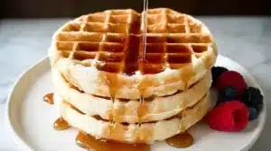 Stack of golden waffles drizzled with maple syrup and served with fresh raspberries and blackberries on a white plate.