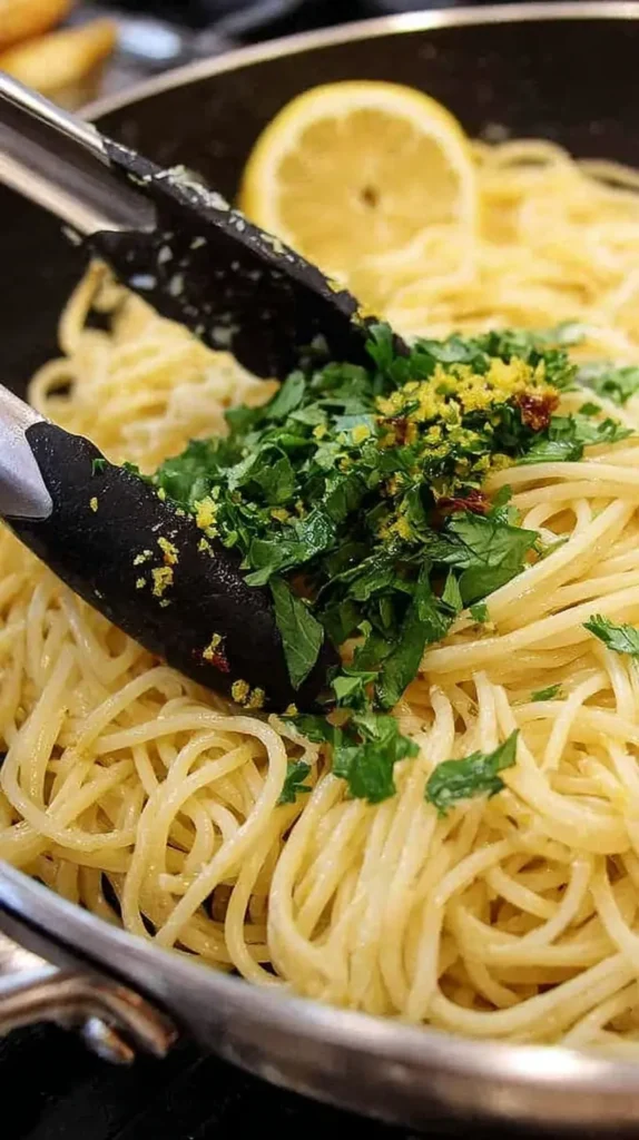 Cooked spaghetti being tossed with fresh parsley and lemon zest in a skillet to finish a bright lemon garlic pasta.