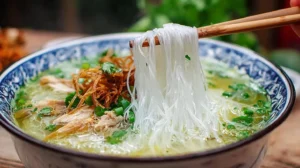 Bowl of Vietnamese chicken noodle soup with clear golden broth; chopsticks lift silky rice noodles over shredded chicken, scallions, cilantro, and crispy fried shallots.