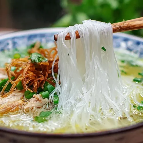 Bowl of Vietnamese chicken noodle soup with clear golden broth; chopsticks lift silky rice noodles over shredded chicken, scallions, cilantro, and crispy fried shallots.