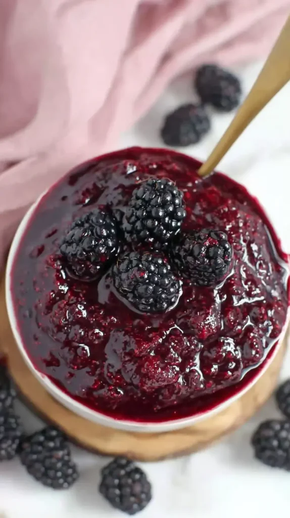 Overhead bowl of glossy blackberry pie filling topped with fresh blackberries, vintage spoon, on a wood board.