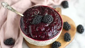 Bowl of glossy blackberry pie filling topped with fresh blackberries, set on a wooden board with scattered berries and a pale linen.
