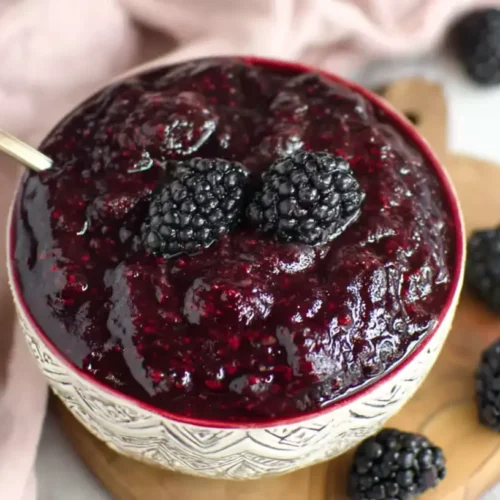 Bowl of glossy blackberry pie filling topped with fresh blackberries, set on a wooden board with scattered berries and a pale linen.