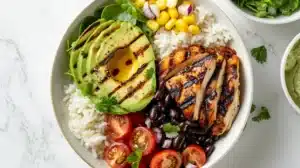 Overhead photo of a chicken avocado bowl with grilled chicken, sliced avocado, white rice, black beans, corn, spinach and cherry tomatoes on a light marble background