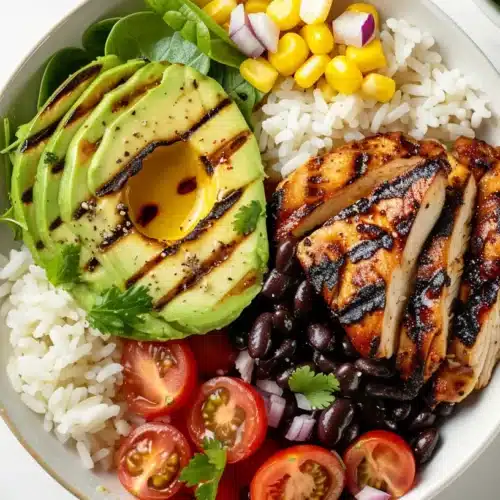 Overhead photo of a chicken avocado bowl with grilled chicken, sliced avocado, white rice, black beans, corn, spinach and cherry tomatoes on a light marble background