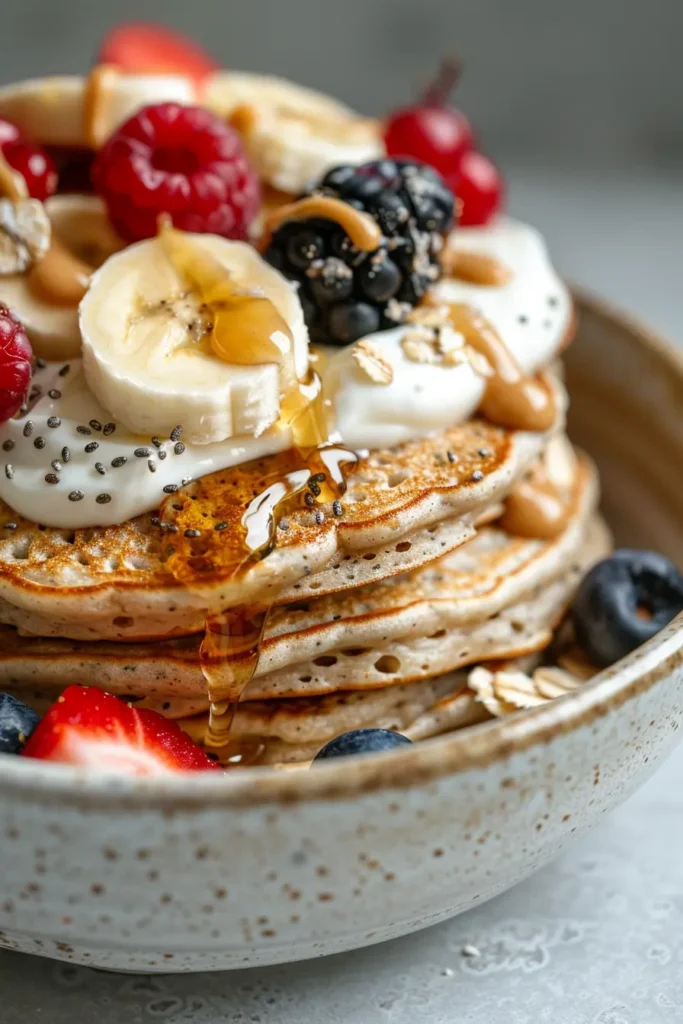 Close-up stack of oat pancakes topped with yogurt, banana slices, berries, chia seeds and honey drizzle in a bowl.