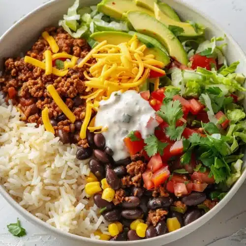 Taco rice bowl with fluffy white rice, seasoned ground beef, black beans, corn, lettuce, avocado, cheddar and creamy sauce in a white bowl