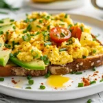 Plate of avocado toast topped with scrambled eggs, chives, and cherry tomatoes.