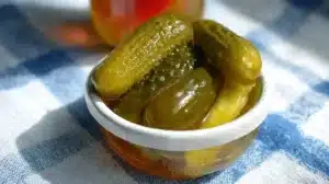 Small white ramekin filled with whole dill pickles sitting in golden brine on a blue-and-white checkered cloth, sunlight highlighting the glossy skins.