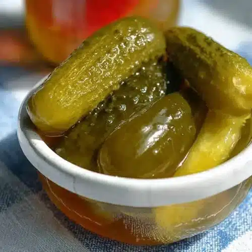 Small white ramekin filled with whole dill pickles sitting in golden brine on a blue-and-white checkered cloth, sunlight highlighting the glossy skins.