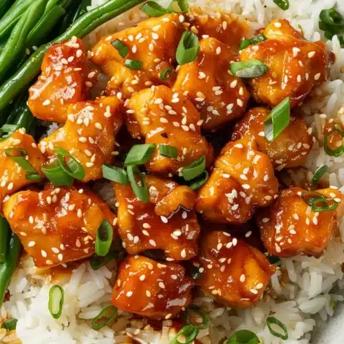 Overhead plate of honey butter chicken bites on rice with a side of green beans and sesame seed garnish.