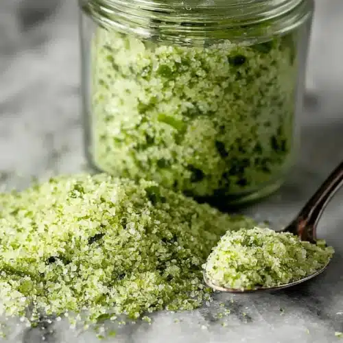 Pile of jalapeño salt spilled on marble with a spoonful next to a small jar.