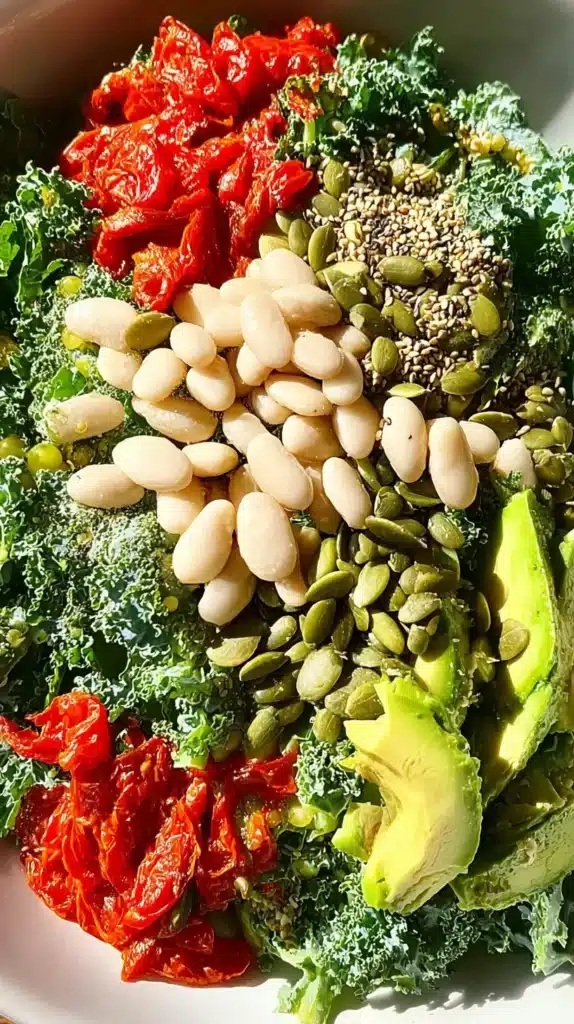 Flat-lay of kale, white beans, avocado slices, pumpkin seeds, and sun-dried tomatoes arranged in a bowl before tossing.