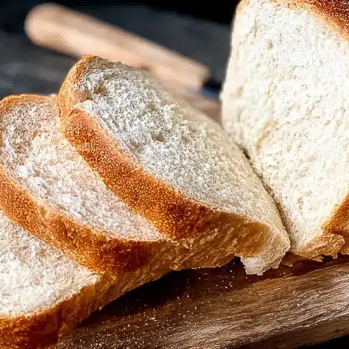 Sliced loaf of soft white bread on a wooden board with a butter dish in the background, made with a potato flake sourdough starter.