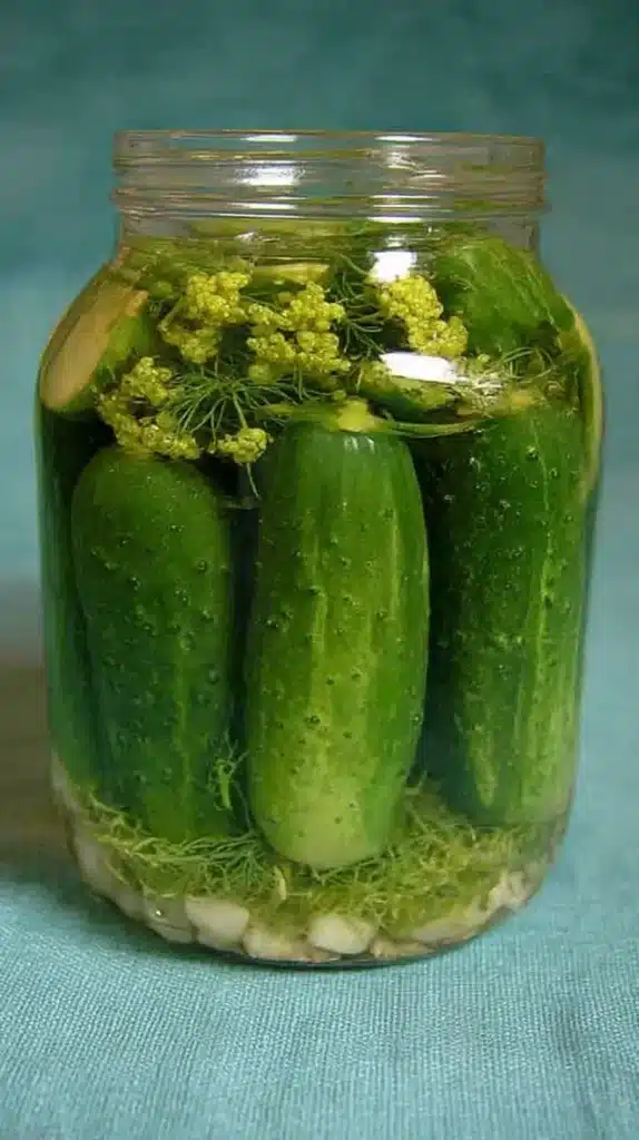 Glass jar packed upright with whole pickling cucumbers, dill blossoms, and minced garlic submerged in clear brine against a teal backdrop.
