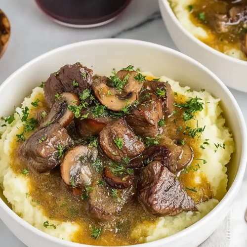 Two bowls of mashed potatoes topped with beef tips and mushrooms, surrounded by chopped herbs and lemon.