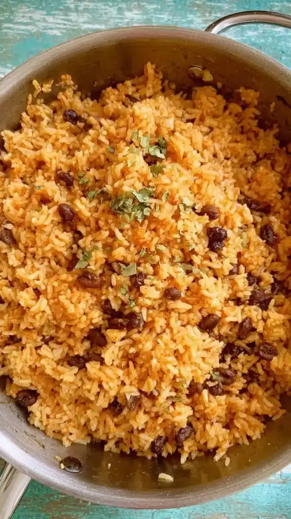 Large stainless steel pot filled with fluffy Puerto Rican rice and black beans, sprinkled with fresh herbs on a teal surface.