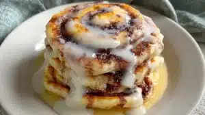 Cinnamon Roll Pancakes: Easy, Fluffy & Wonderful Cinnamon Roll Pancakes stacked on a white plate, swirled with cinnamon sugar and topped with glossy vanilla icing