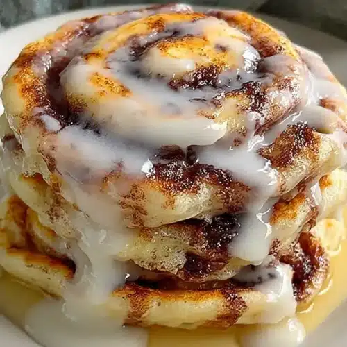 Cinnamon Roll Pancakes: Easy, Fluffy & Wonderful 2 Cinnamon Roll Pancakes stacked on a white plate, swirled with cinnamon sugar and topped with glossy vanilla icing