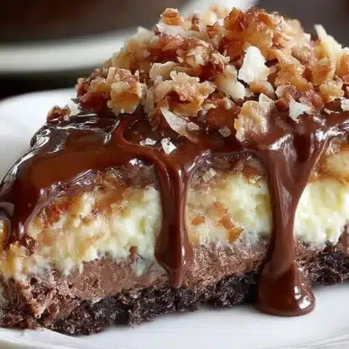 German Chocolate Cheesecake slice on a plate with chocolate ganache topping, coconut-pecan frosting layer, and a dark chocolate cookie crust