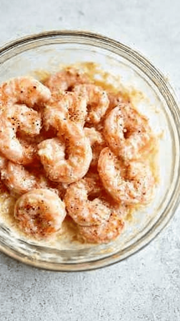 Raw shrimp coated in a creamy seasoned marinade in a clear glass bowl, step one in easy gluten free shrimp