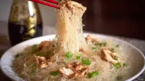 Chopsticks lifting chicken long rice glass noodles with green onions and shredded chicken from a white plate.