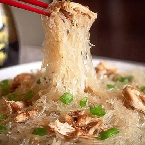 Chopsticks lifting chicken long rice glass noodles with green onions and shredded chicken from a white plate.
