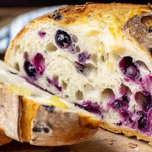 Sliced lemon blueberry sourdough bread on a wooden board, showing a crusty golden crust and airy crumb studded with blueberries.